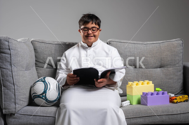 دعم المجتمع السعودي لهوايات أصحاب الهمم, الجلوس بسرور وممارسة هواية المطالعة, الأنشطة الترفيهية المفيدة لذوي الاحتياجات الخاصة, بورتريه لشاب عربي خليجي سعودي مبتسم مصاب بمتلازمة داون يرتدي الثوب التقليدي والنظارة الطبية يقوم بقراءة القصص, خلفية رمادية