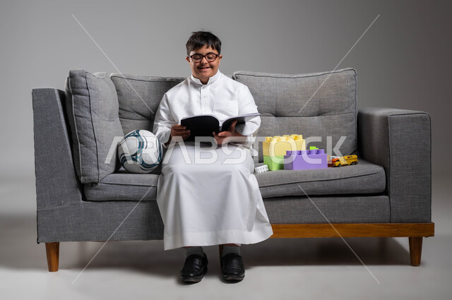 الجلوس بسرور وممارسة هواية المطالعة, الأنشطة الترفيهية المفيدة لذوي الاحتياجات الخاصة, بورتريه لشاب عربي خليجي سعودي مبتسم مصاب بمتلازمة داون يرتدي الثوب التقليدي والنظارة الطبية يقوم بقراءة القصص, دعم المجتمع السعودي لهوايات أصحاب الهمم, خلفية رمادية