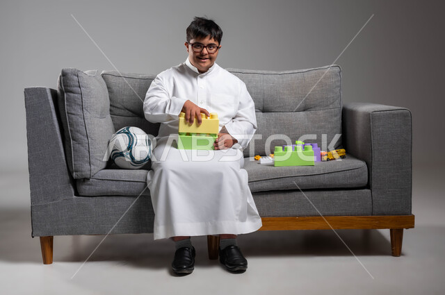 دعم وتنمية القدرات الذهنية والحركية للمعوقين في السعودية, نشاطات ترفيهية لذوي الاحتياجات الخاصة, بورتريه لشاب عربي خليجي سعودي مصاب بمتلازمة داون يرتدي الثوب التقليدي والنظارة الطبية ينظر إلى الكاميرا ويقوم بتركيب قطع المكعبات, خلفية رمادية