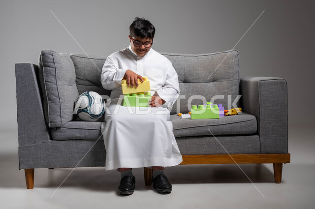 Saudi society’s interest in people of determination, recreational activities for people with special needs, portrait of a Saudi Gulf Arab young man with Down Syndrome wearing a traditional dress and medical glasses assembling cubes, developing mental abilities for people with disabilities, integration into motor activities, gray background