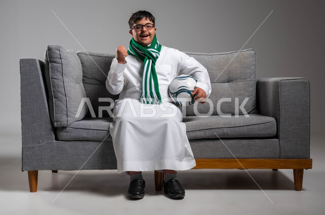 رفع اليد بحماس والسرور لتحقيق الفوز والانتصار, دعم المواهب الرياضية للمعاقين, ارتداء أوشحة تشجيعية للفريق المفضل, بورتريه لشاب عربي خليجي سعودي مبتسم مصاب بمتلازمة داون يرتدي الثوب التقليدي والنظارة الطبية يحمل بيده كرة القدم, خلفية رمادية