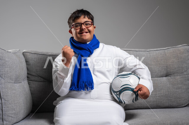 Sitting and raising the hand with gestures of encouragement and enthusiasm, supporting the sports talents of the disabled, portrait of a smiling Gulf Arab Saudi man with Down Syndrome wearing a traditional dress and medical glasses looking at the camera and holding a football in his hand, wearing encouragement scarves for the favorite team, gray background