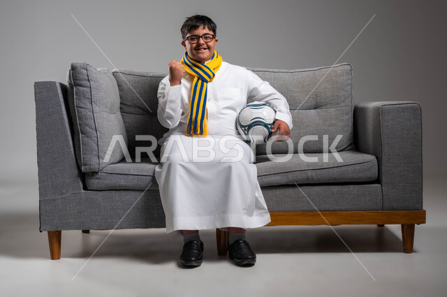 دعم وتنمية المجتمع السعودي للمواهب الرياضية, السعادة والبشاشة بفوز الفريق المفضل, بورتريه لشاب عربي خليجي سعودي مبتسم مصاب بمتلازمة داون يرتدي الثوب التقليدي والنظارة الطبية يحمل بيده كرة القدم, ارتداء أوشحة تشجيعية للفرق الرياضية المفضلة, خلفية رمادية