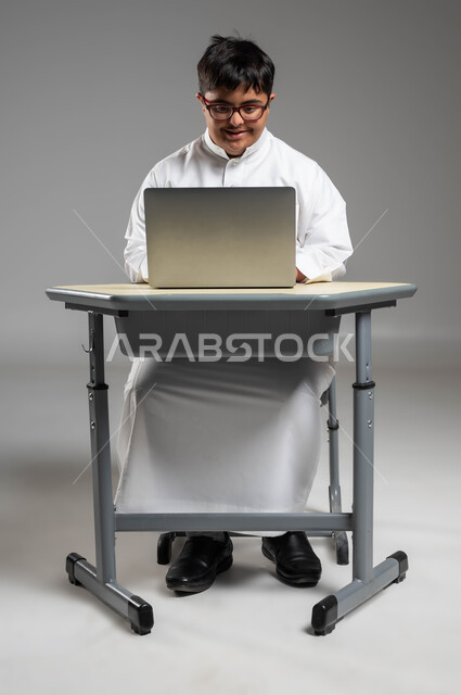 Attention and focus on practical tasks for people of determination, participation of people with special needs in Saudi society, portrait of a smiling Gulf Arab Saudi man with Down Syndrome wearing a traditional dress and medical glasses, using a laptop, gray background