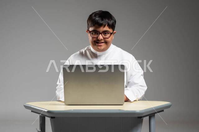 دمج ذوي الاحتياجات الخاصه في الحياه المهنية والعمليه, اهتمام المجتمع السعودي بقدرات أصحاب الهمم, استخدام المعوقين للتقنيات الحديثة, بورتريه لشاب عربي خليجي سعودي مصاب بمتلازمة داون يرتدي الثوب التقليدي والنظارة الطبية يستخدم الحاسب المحمول, خلفية رمادية