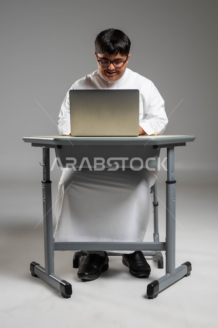 استخدام المعوقين للأجهزة والتقنيات الحديثة, دمج ذوي الاحتياجات الخاصه في الحياه العمليه, بورتريه لشاب عربي خليجي سعودي مبتسم مصاب بمتلازمة داون يرتدي الثوب التقليدي والنظارة الطبية يستخدم الحاسب المحمول, اهتمام المجتمع السعودي بقدرات أصحاب الهمم, خلفية رمادية