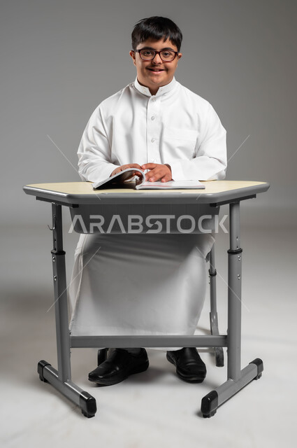 Interest in education and study for people with special needs in Saudi schools, a portrait of a smiling Gulf Arab Saudi man with Down Syndrome wearing a traditional dress looking at the camera, people of determination feeling comfortable and reassured in their environment, gray background