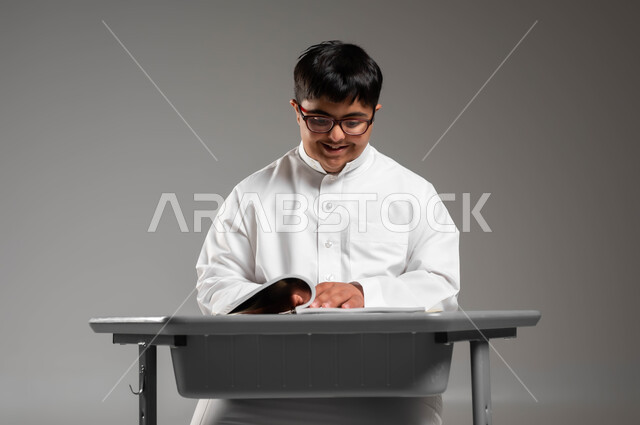 دمج المعاقين في المدارس السعودية, شعور ذوي الهمم بالراحة والطمأنينة في مجتمعهم, بورتريه لشاب عربي خليجي سعودي مبتسم مصاب بمتلازمة داون يرتدي الثوب التقليدي والنظارة الطبية يجلس على المقعد الدراسي, اهتمام المجتمع السعودي بذوي الاحتياجات الخاصة, خلفية رمادية