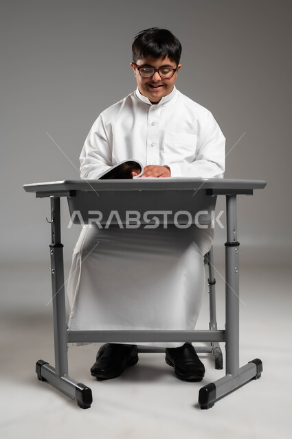 شعور ذوي الهمم بالراحة والطمأنينة في مجتمعهم, بورتريه لشاب عربي خليجي سعودي مبتسم مصاب بمتلازمة داون يرتدي الثوب التقليدي والنظارة الطبية يجلس على المقعد الدراسي, دمج المعاقين في المدارس السعودية, اهتمام المجتمع السعودي بذوي الاحتياجات الخاصة, خلفية رمادية