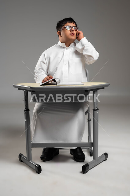 التأمل والنظر إلى شيء ما, اهتمام المجتمع السعودي بتعليم ذوي الاحتياجات الخاصة, دمج المعاقين في المدارس السعوديه, بورتريه لشاب عربي خليجي سعودي مصاب بمتلازمة داون يرتدي الثوب التقليدي والنظارة الطبية يضع يده على خده بإيماءات التفكير, خلفية رمادية