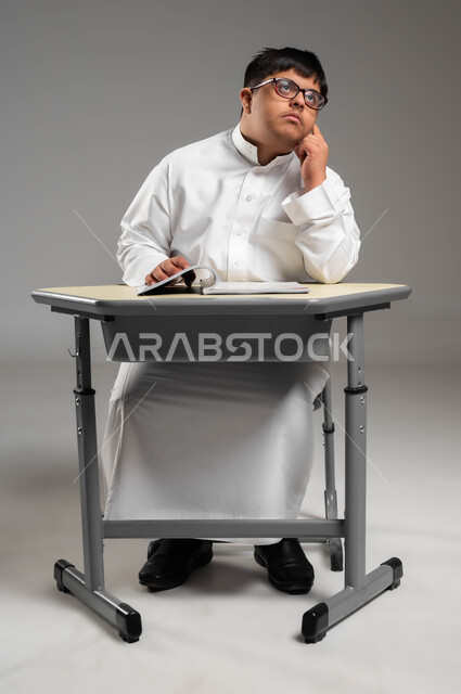 اهتمام المجتمع السعودي بتعليم ذوي الاحتياجات الخاصة, دمج المعاقين في المدارس السعوديه, بورتريه لشاب عربي خليجي سعودي مصاب بمتلازمة داون يرتدي الثوب التقليدي والنظارة الطبية يضع يده على خده بإيماءات التفكير, التأمل والنظر إلى شيء ما, خلفية رمادية