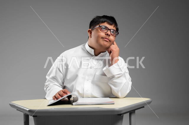 دمج المعاقين في المدارس السعوديه, بورتريه لشاب عربي خليجي سعودي مصاب بمتلازمة داون يرتدي الثوب التقليدي والنظارة الطبية يضع يده على خده بإيماءات التفكير, التأمل والنظر إلى شيء ما, اهتمام المجتمع السعودي بذوي الاحتياجات الخاصة, خلفية رمادية