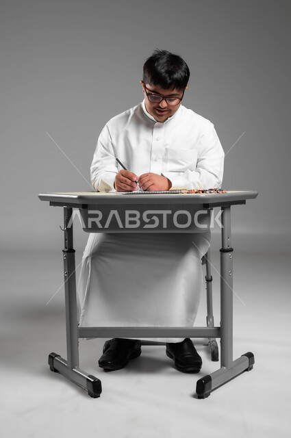 اندماج وتركيز ذوي الهمم بحصة الرسم التعليمية, تنمية المهارة الفنية للمعاقين, دعم المجتمع السعودي لذوي الاحتياجات الخاصة, بورتريه لشاب عربي خليجي سعودي مصاب بمتلازمة داون يرتدي الثوب التقليدي والنظارة الطبية يستخدم أقلام التلوين الخشبية, خلفية رمادية