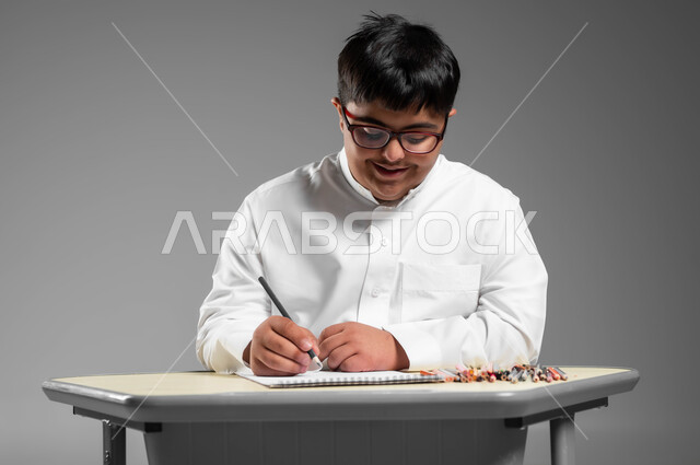 اندماج وتركيز ذوي الهمم  بحصة الرسم التعليمية, تنمية المهارة الفنية للمعاقين, دعم المجتمع السعودي لذوي الاحتياجات الخاصة, بورتريه مقرب لشاب عربي خليجي سعودي مصاب بمتلازمة داون يرتدي الثوب التقليدي والنظارة الطبية يستخدم أقلام التلوين الخشبية, خلفية رمادية