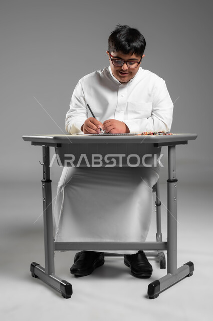 تنمية المهارة الفنية لذوي الهمم, دعم المجتمع السعودي لذوي الاحتياجات الخاصة, استخدام أقلام التلوين الخشبية, بورتريه لشاب عربي خليجي سعودي مصاب بمتلازمة داون يرتدي الثوب التقليدي والنظارة الطبية يقوم بتعلم الرسم والتلوين بتركيز ودقة, خلفية رمادية