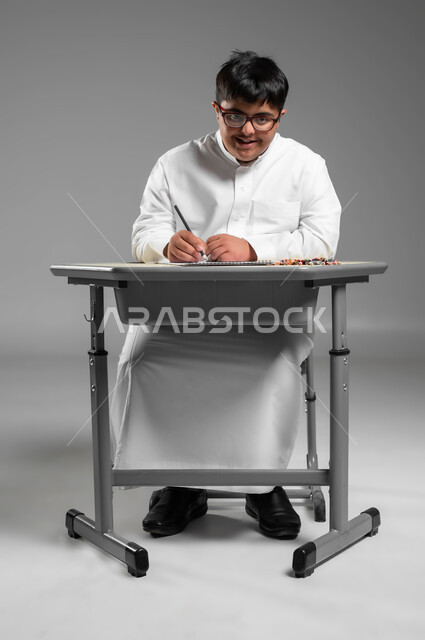 دعم المواهب الفنية لذوي الاحتياجات الخاصة, بورتريه لشاب عربي خليجي سعودي مبتسم مصاب بمتلازمة داون يرتدي الثوب التقليدي والنظارة الطبية يمسك أقلام التلوين الخشبية, دمج المعاقين في المدارس السعودية, الجلوس على المقعد الدراسي والنظر إلى الكاميرا, خلفية رمادية