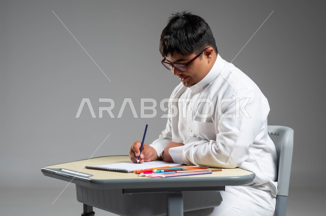 استخدام أقلام التلوين الخشبية, تنمية المهارة الفنية لذوي الهمم, بورتريه لشاب عربي خليجي سعودي مصاب بمتلازمة داون يرتدي الثوب التقليدي والنظارة الطبية يقوم بتعلم الرسم والتلوين بتركيز ودقة, دعم المجتمع السعودي لذوي الاحتياجات الخاصة, خلفية رمادية