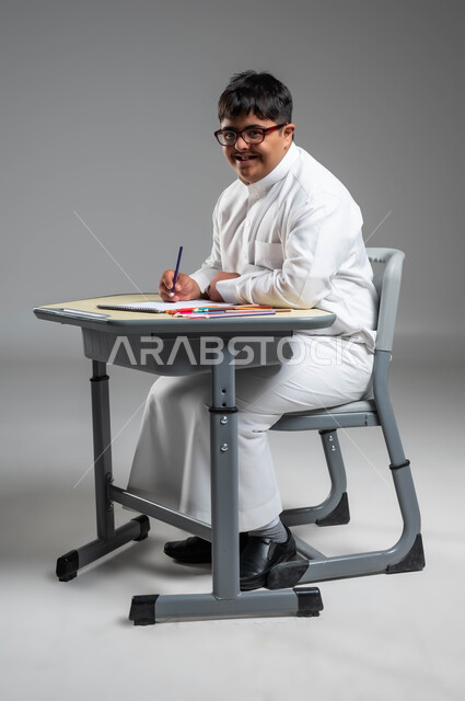 النشاطات الترفيهية لذوي الهمم, تنمية المهارات الفنية للمعاقين, بورتريه لشاب عربي خليجي سعودي مبتسم مصاب بمتلازمة داون يرتدي الثوب التقليدي والنظارة يستخدم أقلام التلوين, اهتمام المجتمع السعودي بالقدرات الحركية والذهنية لذوي الاحتياجات الخاصة, خلفية رمادية