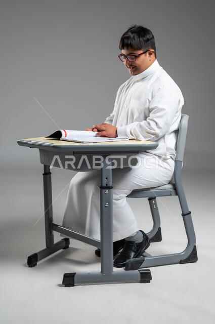 الجلوس على المقعد الدراسي بإيماءات التركيز والاندماج, اهتمام المجتمع السعودي بتعليم ذوي الاحتياجات الخاصة, دمج المعاقين في المدارس السعودية, بورتريه لشاب عربي خليجي سعودي مصاب بمتلازمة داون يرتدي الثوب التقليدي والنظارة الطبية يقوم بقراءة الكتاب, خلفية رمادية
