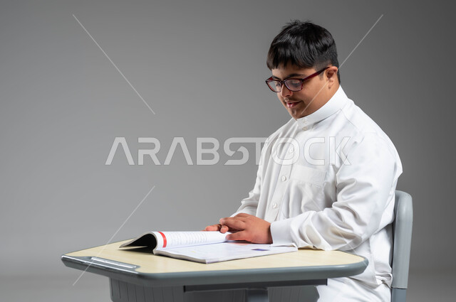 تنمية القدرات العقلية والفكرية لذوي الهمم, اهتمام المجتمع السعودي بتعليم ذوي الاحتياجات الخاصة, دمج المعاقين في المدارس السعودية, بورتريه مقرب لشاب عربي خليجي سعودي مصاب بمتلازمة داون يرتدي الثوب التقليدي والنظارة الطبية يجلس على المقعد الدراسي, خلفية رمادية