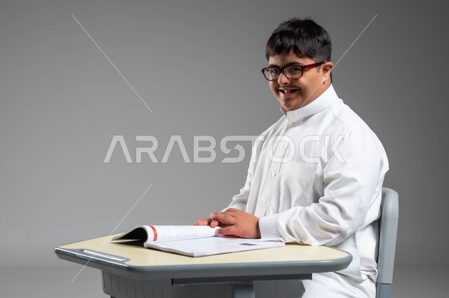 اهتمام المجتمع السعودي بتعليم ذوي الاحتياجات الخاصة, دمج المعاقين في المدارس السعودية, تنمية القدرات العقلية والفكرية لذوي الهمم, بورتريه مقرب لشاب عربي خليجي سعودي مبتسم مصاب بمتلازمة داون يرتدي الثوب التقليدي والنظارة الطبية ينظر إلى الكاميرا, خلفية رمادية