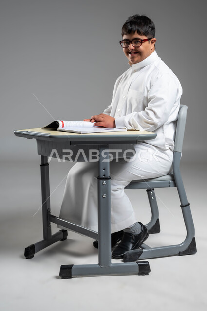Integrating the disabled into Saudi schools, developing the mental and intellectual abilities of people of determination, a portrait of a smiling Gulf Arab Saudi man with Down Syndrome wearing a traditional dress and medical glasses looking at the camera, Saudi society’s interest in educating people with special needs, gray background