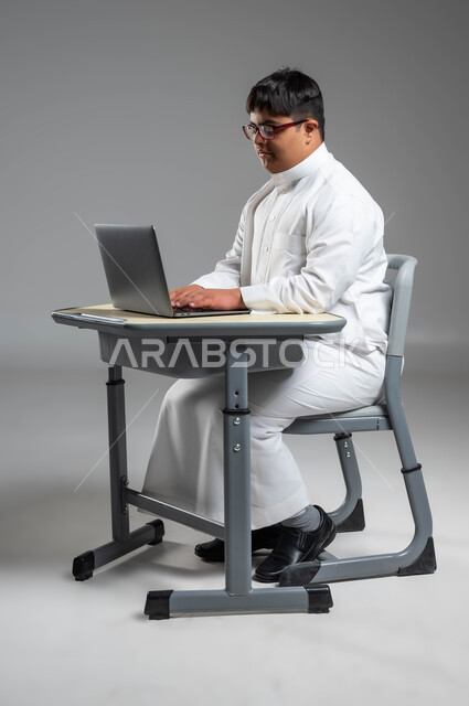 استخدام الأجهزة والتقنيات الحديثة والمتطورة, دمج ذوي الاحتياجات الخاصه في الحياه العمليه, بورتريه لشاب عربي خليجي سعودي مصاب بمتلازمة داون يرتدي الثوب التقليدي والنظارة الطبية يستخدم الحاسب المحمول, اهتمام المجتمع السعودي بقدرات ذوي الهمم, خلفية رمادية