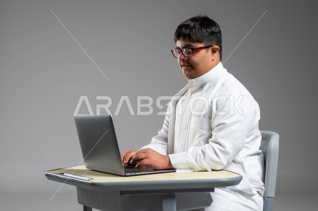 التعليم عن بعد عبرالتقنيات المتطورة, دمج ذوي الاحتياجات الخاصه في الحياه العمليه, بورتريه من الجانب لشاب عربي خليجي سعودي مصاب بمتلازمة داون يرتدي الثوب التقليدي والنظارة الطبية يستخدم الحاسب المحمول, اهتمام المجتمع السعودي بقدرات ذوي الهمم, خلفية رمادية