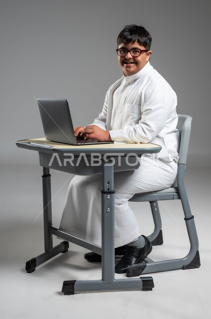 اهتمام المجتمع السعودي بقدرات ذوي الهمم, بورتريه لشاب عربي خليجي سعودي مبتسم مصاب بمتلازمة داون يرتدي الثوب التقليدي والنظارة الطبية ينظر إلى الكاميرا ويستخدم الحاسب المحمول, دمج ذوي الاحتياجات الخاصه في الحياه المهنيه والعمليه, خلفية رمادية