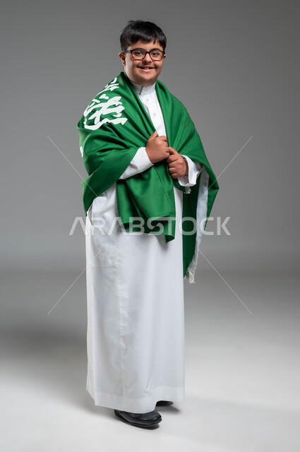 الوقوف بفخر والنظر إلى الكاميرا, مشاركة ذوي الهمم بالأعياد والمناسبات الوطنية, بورتريه لشاب عربي خليجي سعودي مصاب بمتلازمة داون يرتدي الثوب التقليدي والنظارة الطبية يمسك علم المملكة باعتزاز, ذكرى اليوم الوطني في 23 سبتمبر, صورة الجسم بالكامل, خلفية رمادية