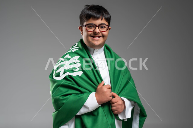استذكار اليوم الوطني السعودي في23 سبتمبر, مشاركة ذوي الهمم بالأعياد والمناسبات الوطنية, التعبير عن حب الوطن والانتماء إليه, بورتريه مقرب لشاب عربي خليجي سعودي مصاب بمتلازمة داون يرتدي الثوب التقليدي والنظارة الطبية يمسك علم المملكة باعتزاز, خلفية رمادية