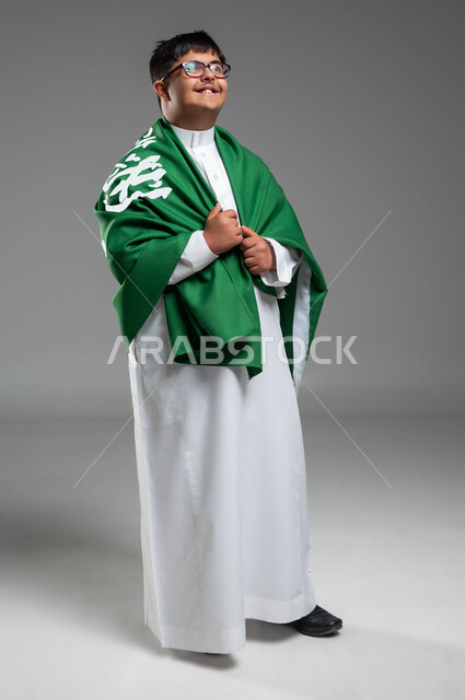 التعبير عن حب الوطن والانتماء إليه, مشاركة ذوي الهمم بالأعياد والمناسبات الوطنية, بورتريه لشاب عربي خليجي سعودي مصاب بمتلازمة داون يرتدي الثوب التقليدي والنظارة الطبية يمسك علم المملكة باعتزاز, ذكرى اليوم الوطني في 23 سبتمبر, صورة الجسم بالكامل, خلفية رمادية