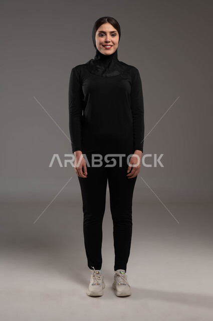 الوقوف باستقامة وانضباط، إيماءات الثقة والاعتزاز بالنفس، الاهتمام بالمظهر الخارجي، بورتريه لفتاة عربية خليجية سعودية ترتدي ملابس الرياضة والحجاب الأسود تقوم بالنظر باتجاه الكاميرا بتعابير الفرح، أنشطة رياضية في السعودية، صورة الجسم بالطول الكامل، خلفية رمادية