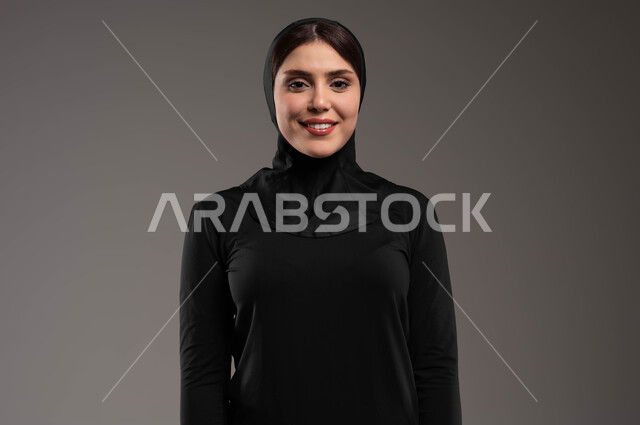 الاهتمام بالمظهر الخارجي، بورتريه مقرب لفتاة عربية خليجية سعودية ترتدي ملابس الرياضة والحجاب الأسود تقوم بالنظر باتجاه الكاميرا بتعابير الفرح، إيماءات الثقة والاعتزاز بالنفس، نادي رياضي نسائي، أنشطة رياضية في السعودية، خلفية رمادية