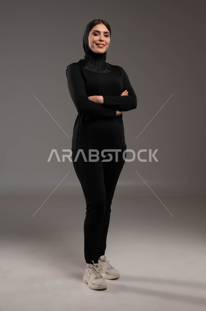 إيماءات الثقة بالنفس والاعتزاز، الاهتمام بالمظهر الخارجي، مفهوم الانوثة والاناقة والنعومة، بورتريه لفتاة عربية خليجية سعودية محجبة ترتدي ملابس الرياضة تقف باستقامة وتنظر إلى كاميرا بحركات أيدي متقاطعة، نادي رياضي نسائي، صورة الجسم بالطول الكامل، خلفية رمادية