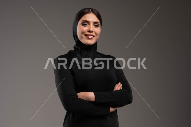 نادي رياضي نسائي، الاهتمام بالمظهر الخارجي، إيماءات الثقة بالنفس والاعتزاز، مفهوم الانوثة والاناقة والنعومة، بورتريه مقرب لفتاة عربية خليجية سعودية محجبة ترتدي ملابس الرياضة تقف باستقامة وتنظر إلى كاميرا بحركات أيدي متقاطعة، خلفية رمادية