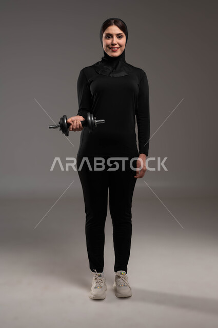 الوقوف باستقامة والاستعداد لرفع الأثقال، كمال الأجسام وتقوية وبناء العضلات، بورتريه لفتاة عربية خليجية سعودية ترتدي ملابس الرياضة والحجاب الأسود، نشاط بدني للمحافظة على الصحة واللياقة الجسدية بايماءات الحماس والسرور، صورة الجسم بالطول الكامل، خلفية رمادية