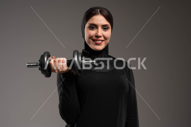 رفع الأوزان الحديدية، تقوية وبناء العضلات، بورتريه مقرب لفتاة عربية خليجية سعودية تقوم بممارسة التمارين الرياضية مع الدامبل، أنشطة رياضية نسائية في السعودية، الحفاظ على الصحة واللياقة الجسدية، ممارسة تمارين رفع الاوزان، خلفية رمادية