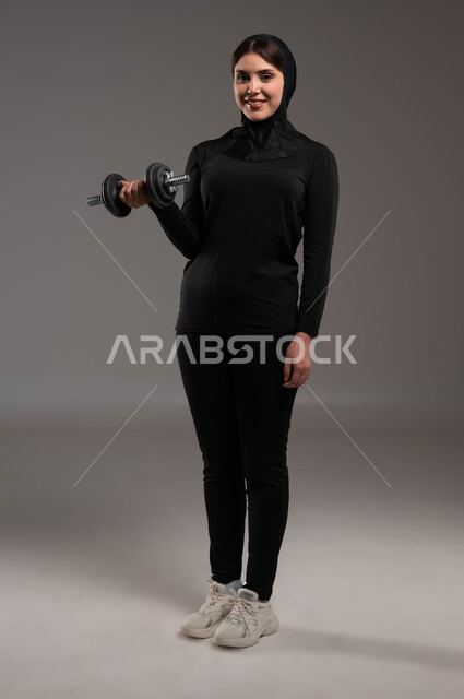 القوة والتحفيز وبناء العضل، ممارسة التمارين الرياضية مع الدمبل، نشاط بدني للمحافظة على الصحة واللياقة الجسدية، بورتريه لفتاة عربية خليجية سعودية ترتدي الحجاب وملابس الرياضة تقوم بتمارين رفع الأثقال (دمبلز)، حرق الدهون والسعرات الحراريه, صورة الجسم بالطول الكامل, خلفية رمادية