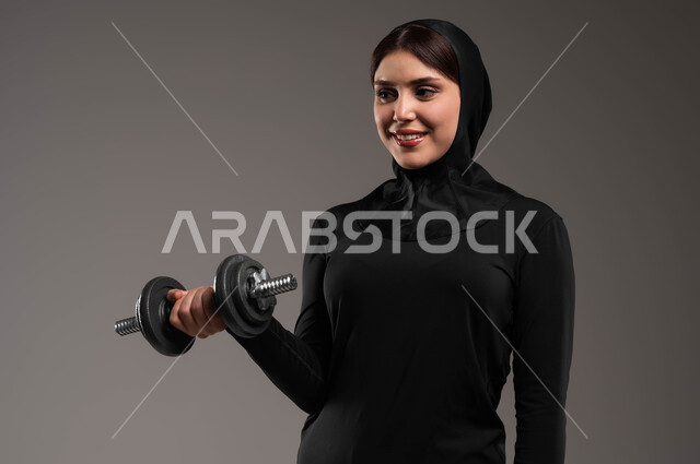 ايماءات الحماس والفرح والاستعداد لممارسة التمارين الرياضية، حمل الأثقال نشاط رياضي للمحافظة على صحة الجسم،  بورتريه مقرب لفتاة عربية خليجية سعودية ترتدي الحجاب وملابس الرياضة تقوم بتمارين رفع الأثقال (دمبلز)، حرق الدهون والسعرات الحراريه,  خلفية رمادية