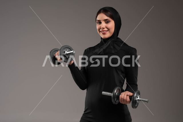 حمل الأثقال نشاط رياضي للمحافظة على صحة الجسم، حرق الدهون والسعرات الحراريه, بورتريه مقرب لفتاة عربية خليجية سعودية ترتدي الحجاب وملابس الرياضة تقوم بتمارين رفع الأثقال (دمبلز)، ايماءات الحماس والفرح والاستعداد لممارسة التمارين الرياضية، خلفية رمادية
