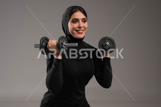 رياضة كمال الأجسام، تمارين رفع الأثقال (دمبلز)،  نشاط بدني للمحافظة على الصحة  اللياقة الجسدية، بورتريه مقرب لفتاة عربية خليجية سعودية تقوم بممارسة التمارين الرياضية مع الدمبل، تقوية عضلات الجسم و بنائها، نادي رياضي نسائي، خلفية رمادية