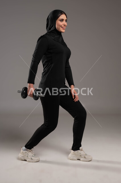 تدريبات رفع الأوزان الحديديه الثقيله، بورتريه من الجانب لفتاة عربية خليجية سعودية محجبة ترتدي الملابس الرياضية, نشاط بدني للمحافظه على الصحه واللياقه الجسديه, تمارين رفع الأثقال (دمبلز), بناء جسم صحي, حرق الدهون والسعرات الحراريه، صورة الجسم بالطول الكامل, خلفية رمادية