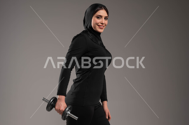 Strength, challenge and motivation for a healthy lifestyle, strengthening and building body muscles, weight lifting exercises, close-up portrait of a veiled Saudi Gulf Arab girl, exercising with dumbbells, physical activity to maintain health and physical fitness, lifting heavy iron weights