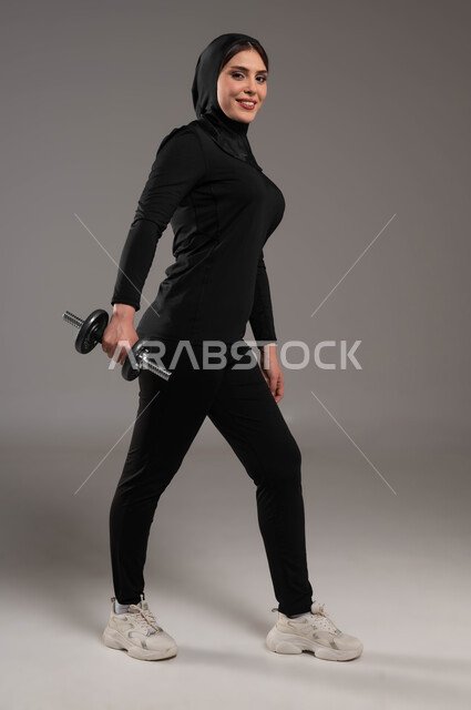 تمارين حمل الاوزان والاثقال، خسارة السعرات الحرارية، بورتريه لفتاة عربية خليجية سعودية محجبة تمارس الرياضة بهمة وقوة، ايماءات الحماس والسرور، نشاط بدني للمحافظة على الصحة و اللياقة الجسدية ، تقوية عضلات الجسم و بنائها، نادي رياضي نسائي، صورة الجسم بالطول الكامل,خلفية رمادية