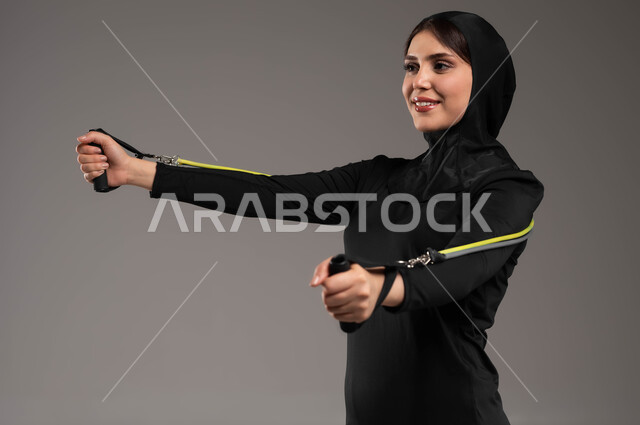 تمارين اليدين والصدر المتوسع، نشاط بدني للمحافظة على الصحة واللياقة الجسدية، تمارين الحبل المطاطي، نادي رياضي نسائي، ممارسة التمارين الرياضية مع قبضة اليد المطاطية،  بورتريه مقرب لفتاة عربية خليجية سعودية محجبة، ترتدي الملابس الرياضية، أسلوب حياة صحي, خلفية رمادية