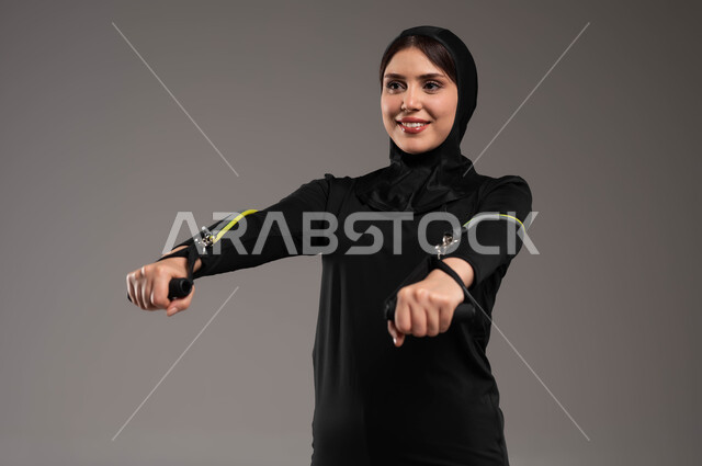 ممارسة التمارين الرياضية مع قبضة اليد المطاطية،  بورتريه مقرب لفتاة عربية خليجية سعودية محجبة، ترتدي الملابس الرياضية، أسلوب حياة صحي، تمارين اليدين والصدر المتوسع، نشاط بدني للمحافظة على الصحة واللياقة الجسدية، تمارين الحبل المطاطي, خلفية رمادية