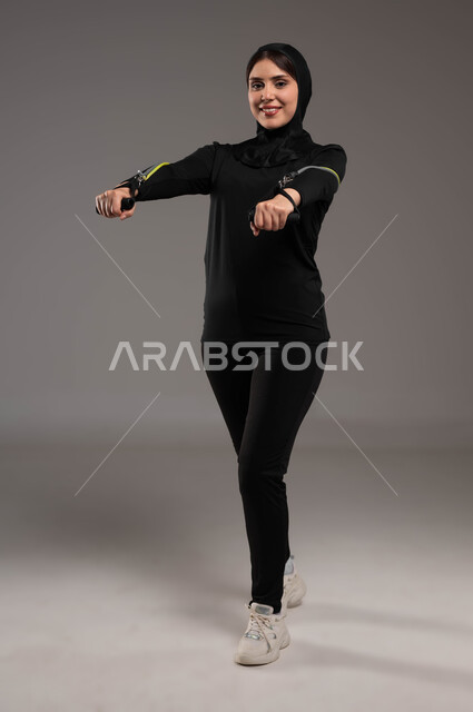 نشاط بدني للحفاظ على الصحة واللياقة الجسدية، تمارين اليدين والصدرالمتوسع وحبل المقاومة، محاولة انقاص الوزن، بورتريه لشابة عربية خليجية سعودية محجبة تقوم بالتمرين مع قبضة اليد المطاطية، أدوات ومعدات رياضية، أسلوب حياة صحي، صورة لكامل الجسم، خلفية رمادية