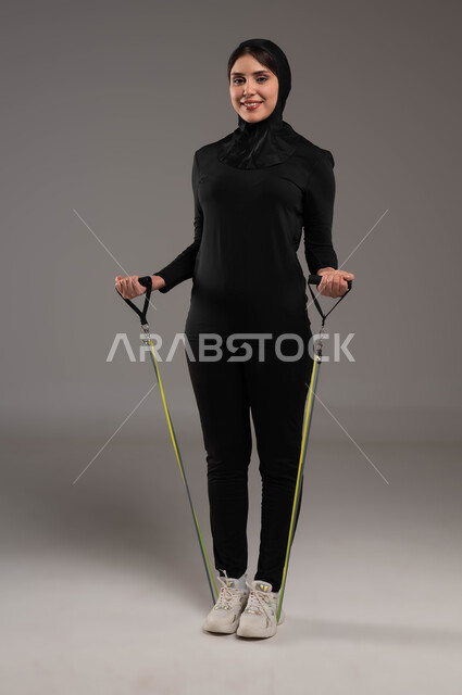 تمارين اليدين والصدرالمتوسع وحبل المقاومة، محاولة انقاص الوزن، بورتريه لشابة عربية خليجية سعودية محجبة تقوم بالتمرين مع قبضة اليد المطاطية، نشاط بدني للمحافظة على الصحة واللياقة الجسدية، أدوات ومعدات رياضية، أسلوب حياة صحي، صورة لكامل الجسم، خلفية رمادية