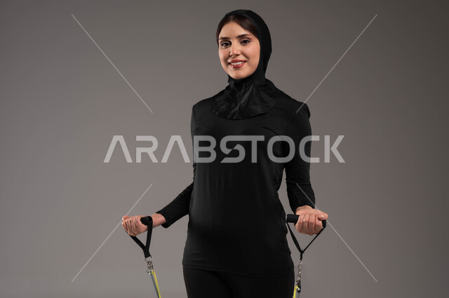 تمارين حبل المقاومة لحرق السعرات الحرارية، التمرن مع قبضة اليد المطاطية و المحافظةعلى أسلوب حياة صحي، تقوية عضلات الجسم والحفاظ على اللياقة، نشاط بدني للعناية بالصحة الجسدية، محاولة انقاص الوزن، بورتريه مقرب لشابة عربية خليجية سعودية محجبة، خلفية رمادية